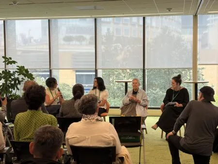 Panel of people seated in a circle talking in a bright meeting room with large windows and blinds behind them.