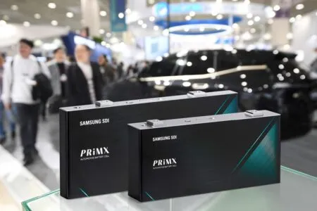 Two Samsung SDI PriMX automotive battery cells on a glass display at a busy expo, with a car in the blurred background.