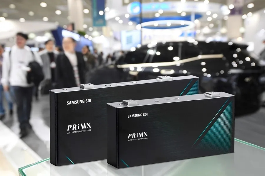 Two Samsung SDI PriMX automotive battery cells on a glass display at a busy expo, with a car in the blurred background.