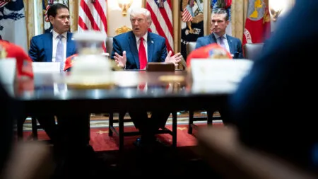Donald Trump speaks at a formal meeting, flanked by two aides, with U.S. flags behind him in a gold-toned room.