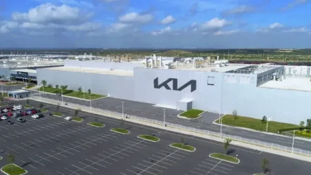 Aerial view of a large white Kia building with an expansive parking lot and a blue sky above.