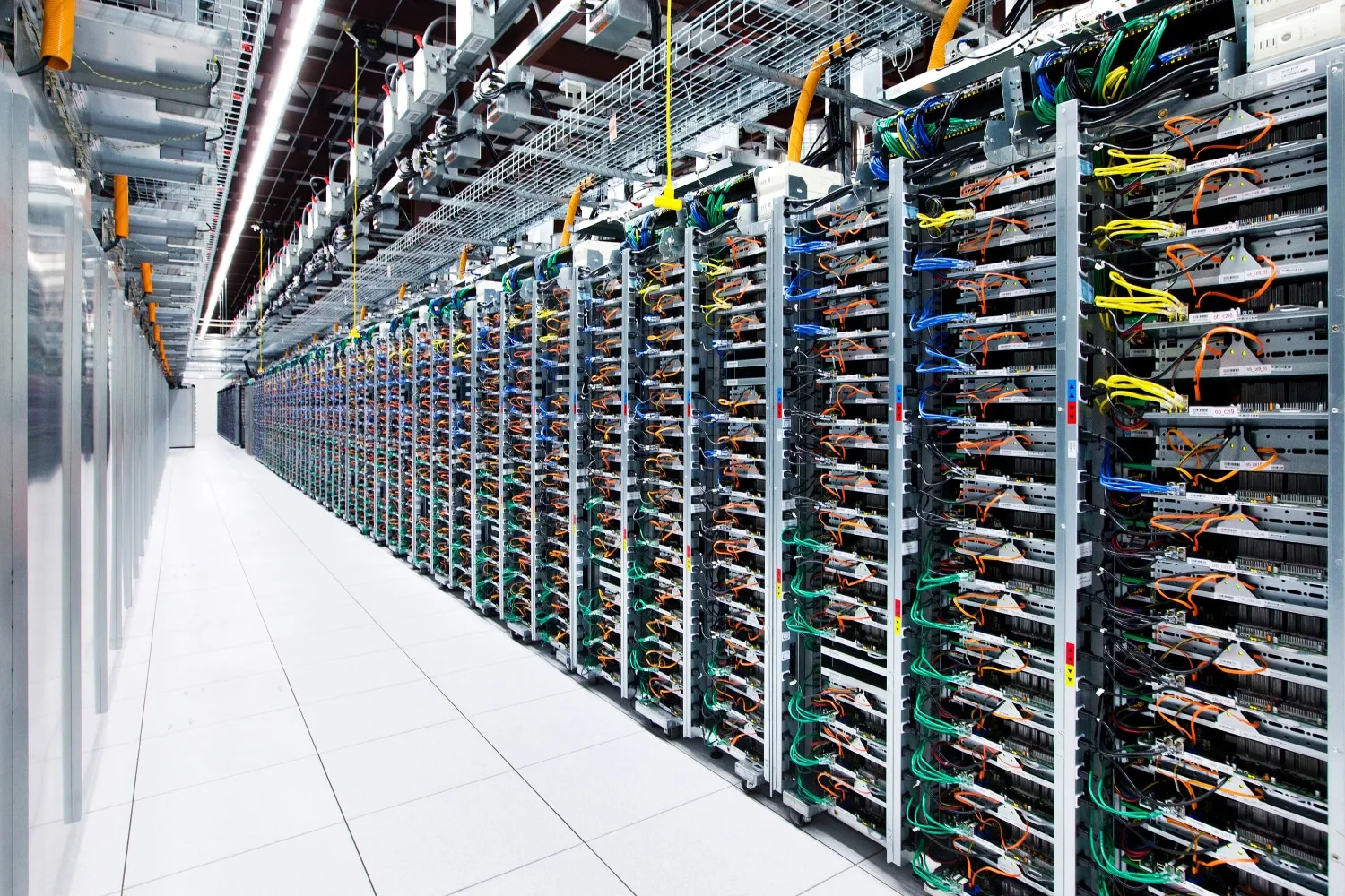Long corridor of data center server racks with many multicolored cables and overhead wiring