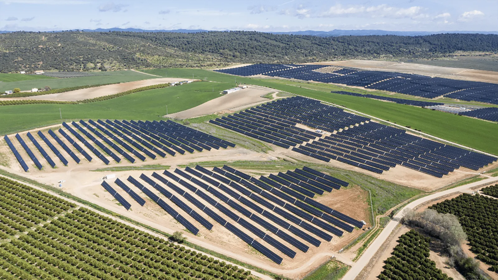 Aerial view of a solar farm with rows of dark solar panels across rolling fields, under a blue sky.