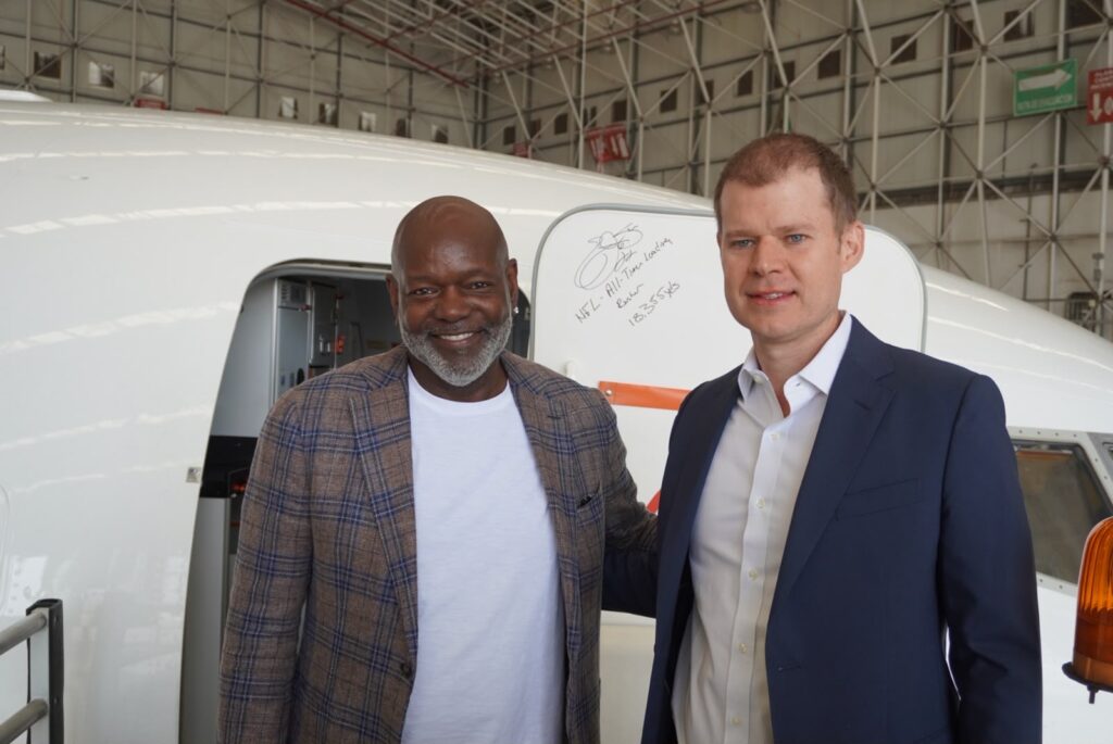 - Petróleo y Energía Two men pose beside a small white airplane inside a hangar; one wears a plaid blazer and white shirt, the other a navy suit, both smiling.