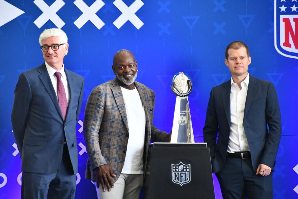 AEROMEXICO 2 - Petróleo y Energía Three men in suits pose with the NFL Lombardi Trophy on a blue stage backdrop.