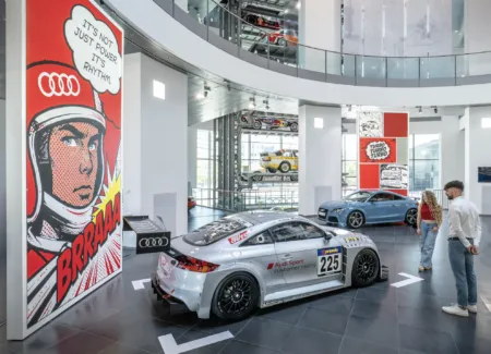 Audi showroom with race cars on display, large pop-art poster on left and visitors in foreground browsing exhibits
