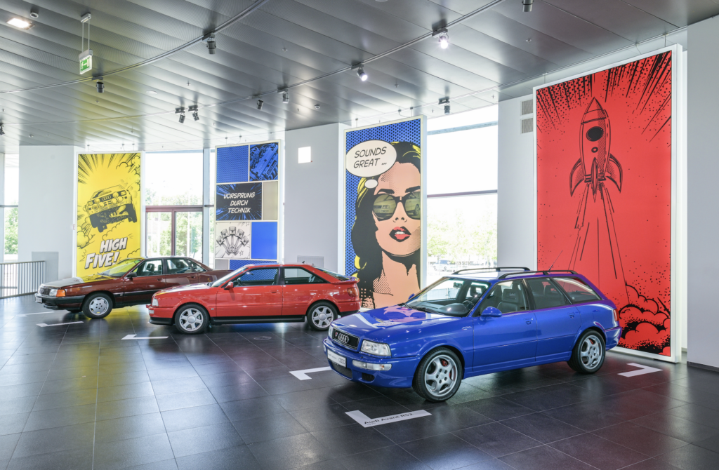 AUDI MUSEO 2026 2 - Petróleo y Energía Showroom with comic-style wall panels and several Audi cars, including red and blue models, indoors.