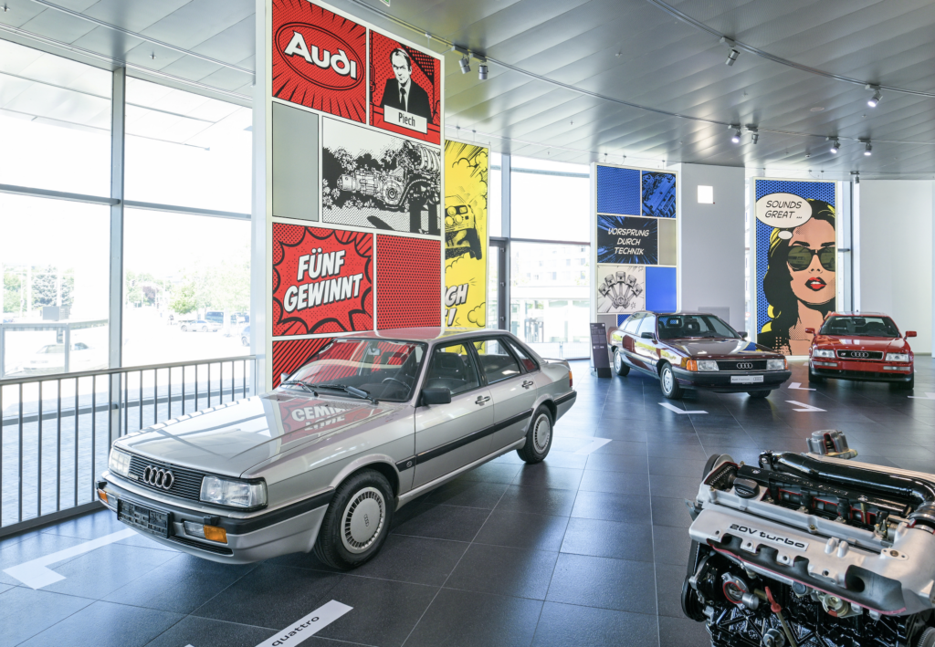 AUDI MUSEO 2026 3 - Petróleo y Energía Inside an Audi showroom with silver cars on a dark tiled floor and large pop-art panels along the wall.
