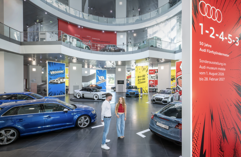 AUDI MUSEO 2026 4 - Petróleo y Energía Couple talking in a bright Audi showroom with multiple cars on display and a bold red display wall on the right.