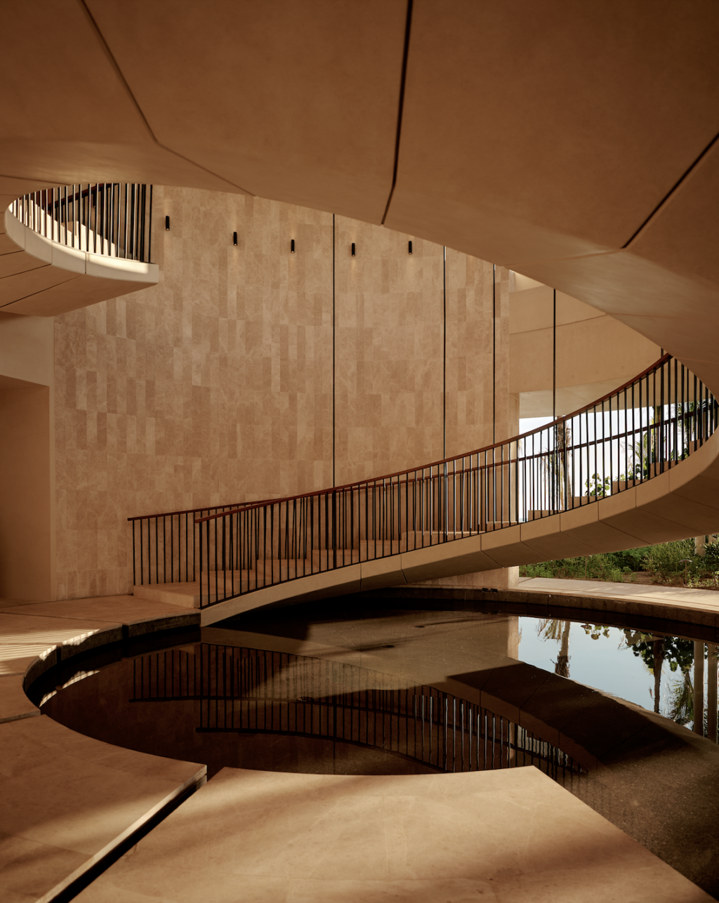 Amanvari 4 - Petróleo y Energía Curved indoor staircase with a black railing looping around a circular reflecting pool and beige stone walls in a modern atrium.