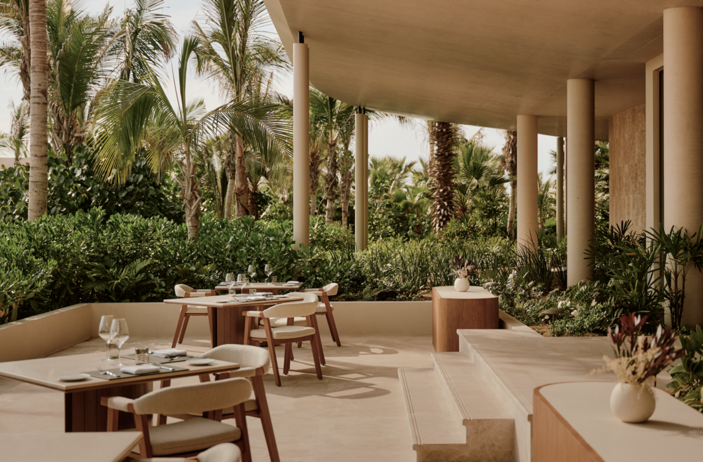 Amanvari 6 - Petróleo y Energía Outdoor dining area on a terrace with wooden tables and chairs, glassware, and lush tropical plants surrounding beige columns under a curved roof.