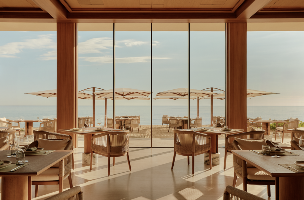 Amanvari 7 - Petróleo y Energía Indoor beachside restaurant with large glass windows and wooden beams, sunlit tables, and view of umbrellas on the sandy beach