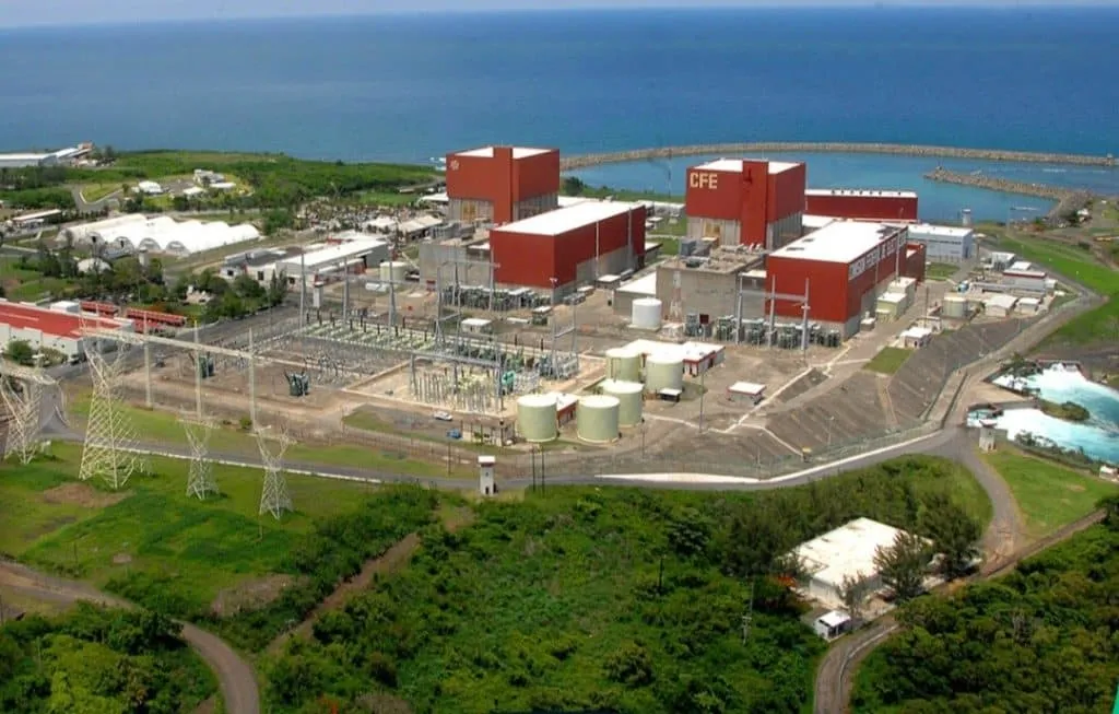 Coastal power plant with red CFE buildings, storage tanks, and electrical equipment near the ocean.