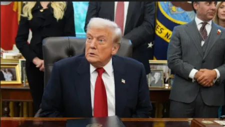 Man in a dark suit with red tie sits at a large desk in an office, with people and portraits in the background.