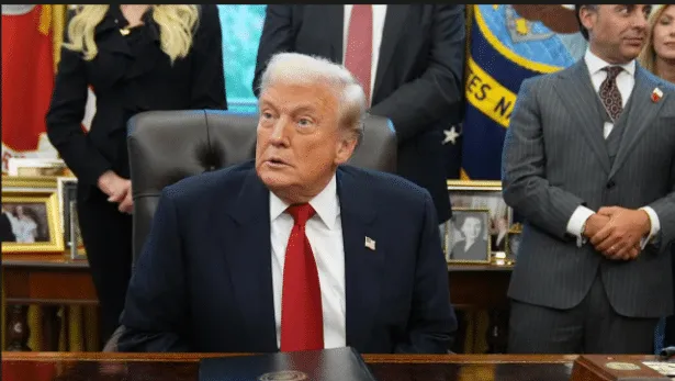 Man in a dark suit with red tie sits at a large desk in an office, with people and portraits in the background.