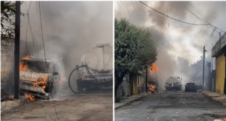 Two vehicles on a smoky street are on fire, with flames and thick smoke rising near utility poles and a tanker truck behind.