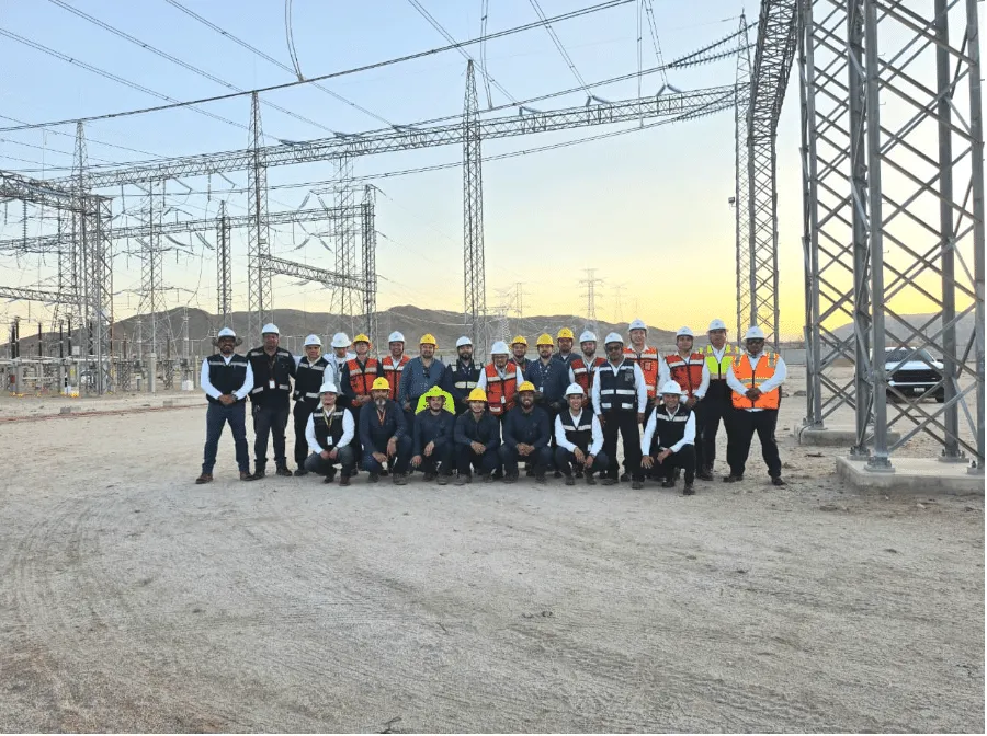 Group of construction workers in hard hats and safety vests posing at an electrical substation site at dusk.