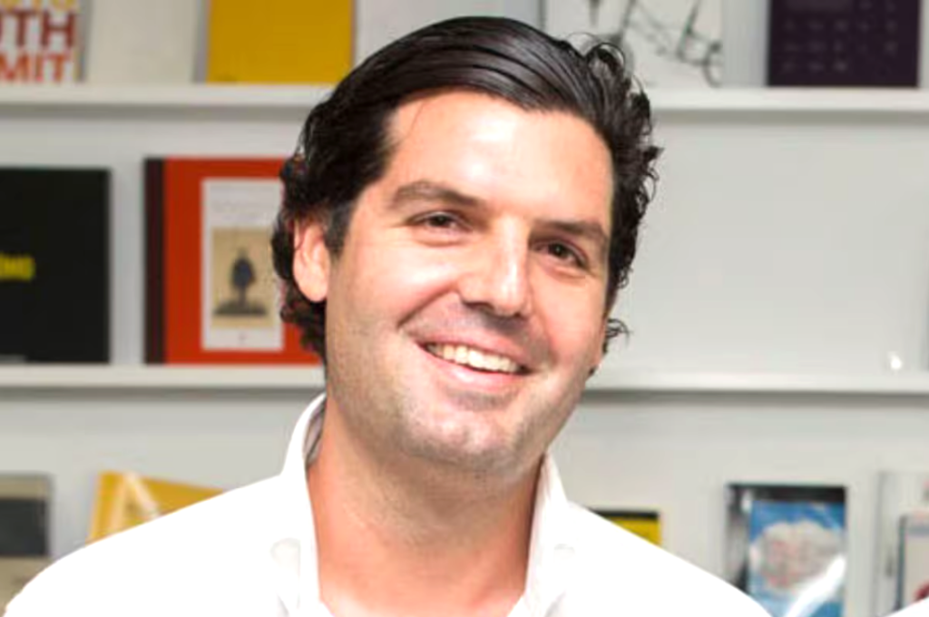 Captura de pantalla 2026-08-18 113607 - Petróleo y Energía Smiling man with dark hair in a white shirt, standing in front of shelves with colorful books or magazines behind him in a bookstore or office.