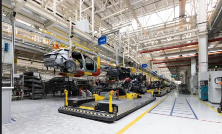 Automotive assembly plant with partially assembled cars on a production line and overhead machinery in a bright industrial space.