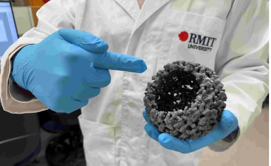 Scientist in a lab coat and blue gloves holding a porous gray ball and pointing at it for scale or emphasis.