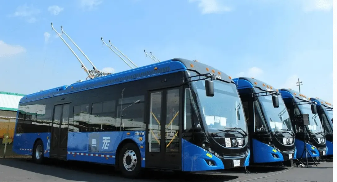 Several blue electric trolleybuses lined up at a depot under a clear sky, with overhead trolley poles visible on top.