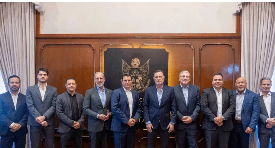 Twelve men in business suits stand shoulder to shoulder in a wood-paneled room with a crest emblem behind them.