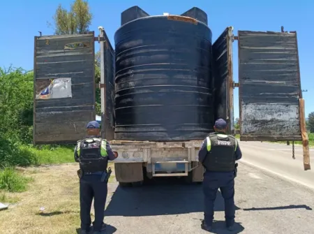 Two officers in dark uniforms inspect a large black cylindrical water tank on a flatbed trailer by the roadside.