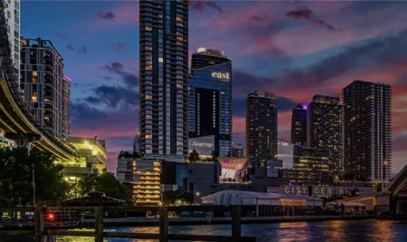Evening city skyline along a waterfront with tall glass buildings and colorful lights, seen from a wooden pier.