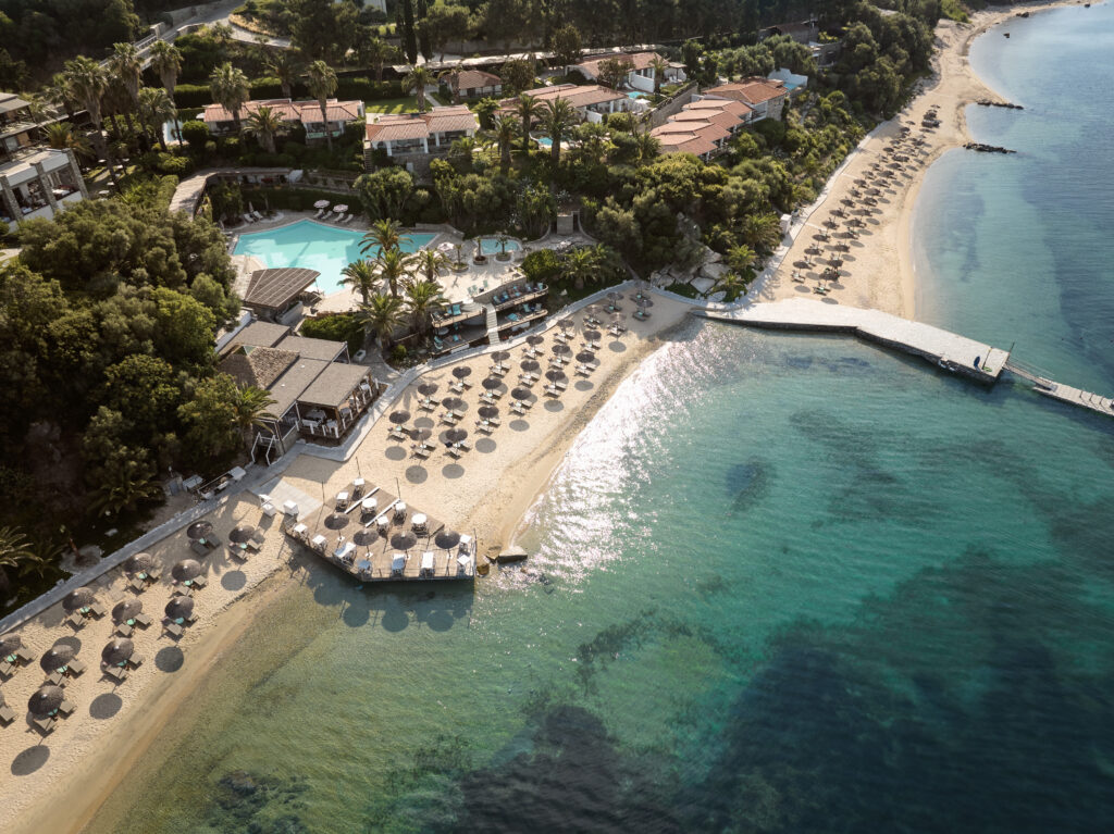 Eagles Palace Exterior - Petróleo y Energía Aerial view of a beach resort with rows of umbrellas along the shore and turquoise water beyond the beach, palm trees and villas in the background.