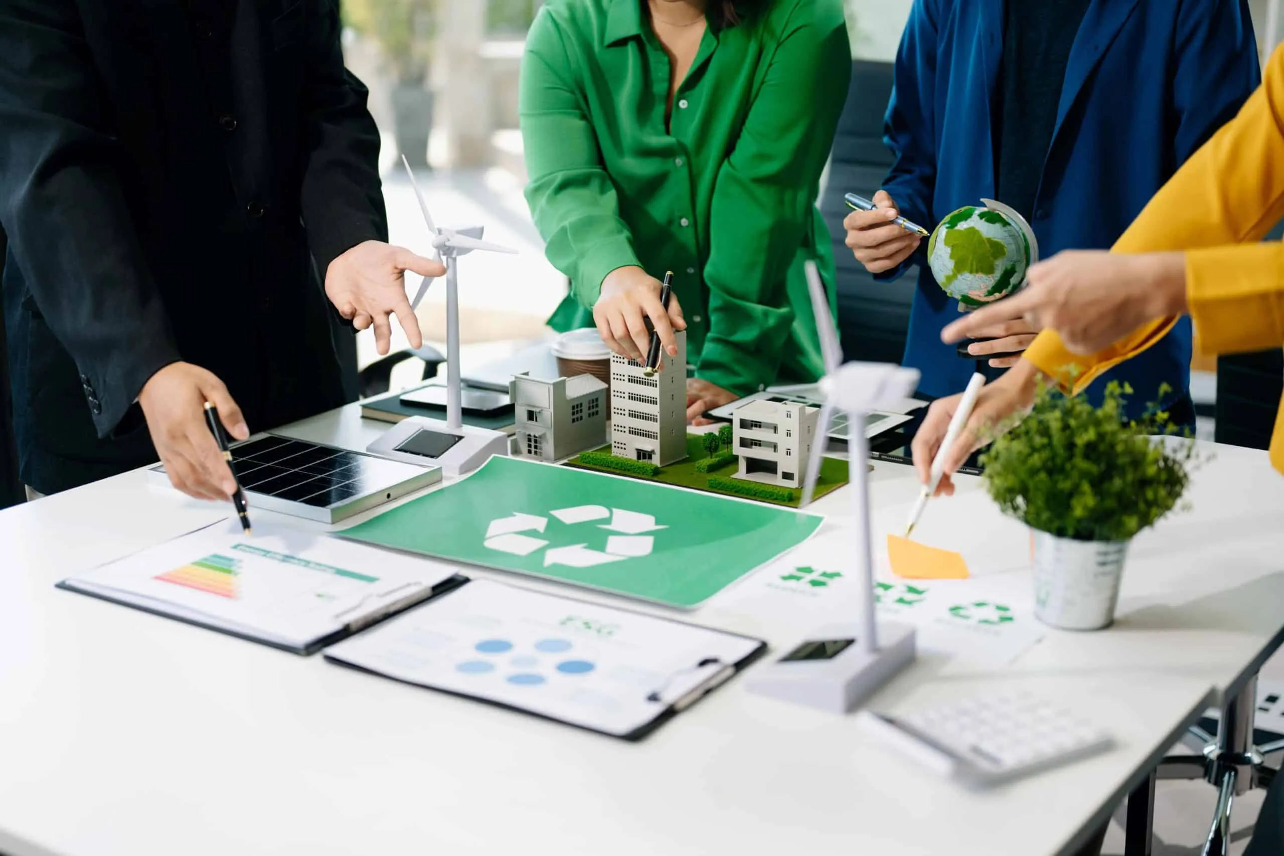 Team of colleagues collaborates over architectural models and green plan on a conference table, featuring a wind turbine and plants for sustainability.