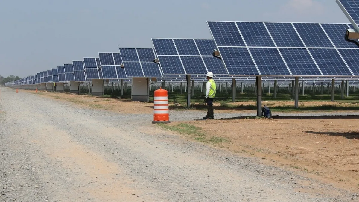 Solar farm with rows of panels; a construction worker in a safety vest and hard hat stands near an orange traffic barrel.