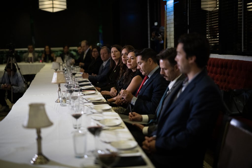 FLA_2488 - Petróleo y Energía Long banquet table with formally dressed attendees seated along one side during a dimly lit event, plates and glasses set before them.