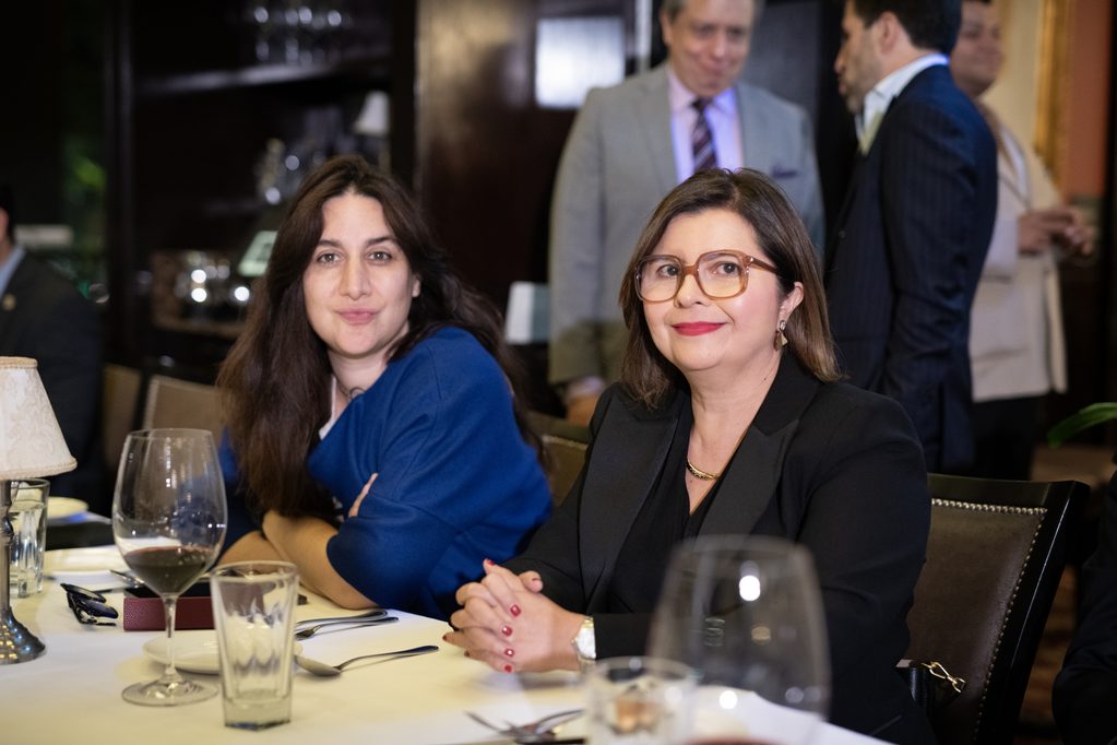 FLA_2579 - Petróleo y Energía Two women sit at a formal dinner table with wine glasses and plates, smiling at the camera in a restaurant setting.