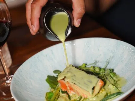 Person pours pale green sauce from a small pitcher onto a plated salmon with greens in a light blue bowl.