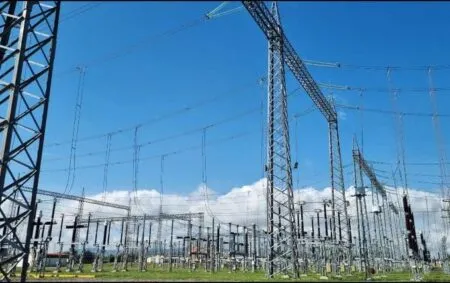 High-voltage power substation with steel lattice towers, wires, and transformers under a blue sky.