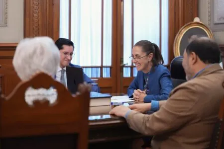 Smiling woman in blue blazer discusses documents with colleagues around a wooden conference table in a formal meeting room.