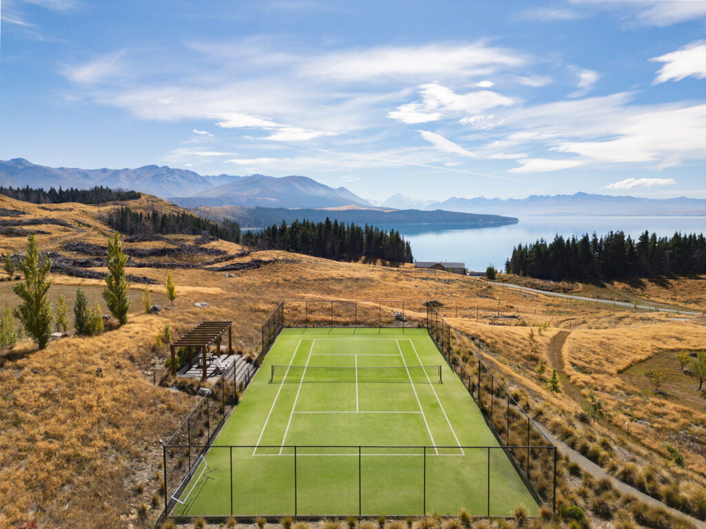 - Petróleo y Energía Empty outdoor tennis court fenced in, with golden hills, trees, a lake, and distant mountains under a blue sky.