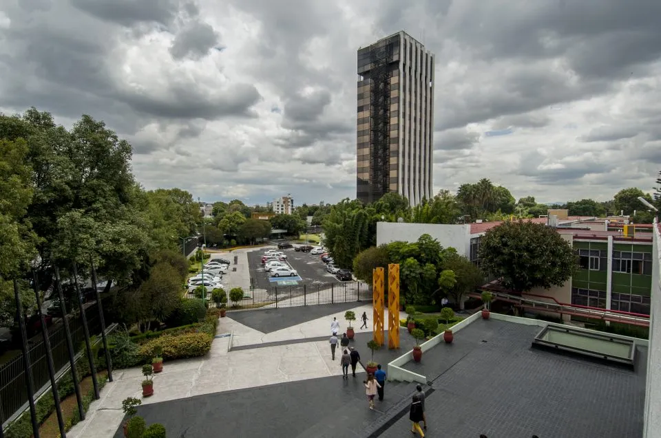 Aerial view of a city plaza with a tall striped high-rise, trees, and people walking toward the building. A parking lot is visible in the background behind a fence.