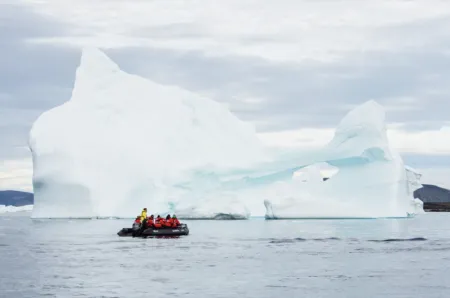 Inflatable raft with people wearing orange life jackets near a massive iceberg in icy blue water under a cloudy sky.