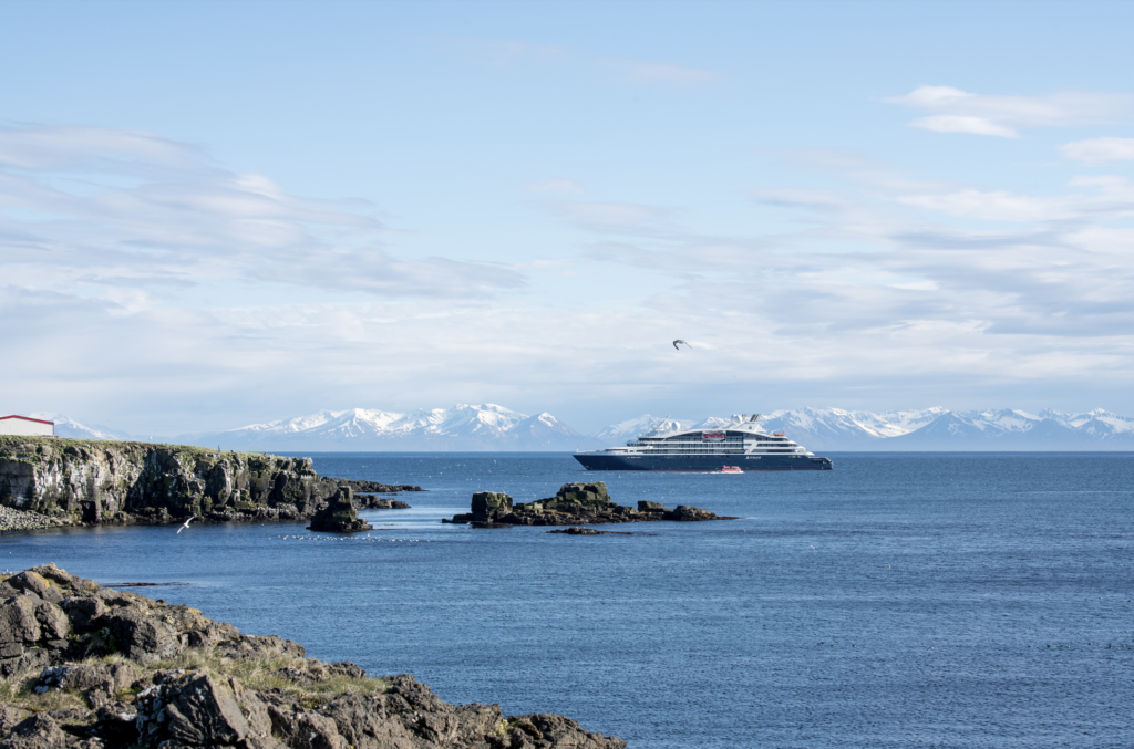 - Petróleo y Energía Cruise ship docked off a rocky coastline with snow-covered mountains in the distance under a blue sky.