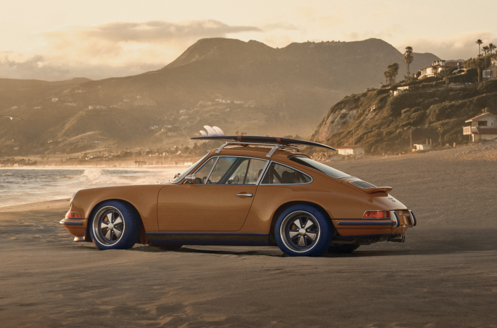 LV_4 - Petróleo y Energía Orange vintage car parked on a sandy beach at sunset, with a surfboard on its roof and distant hills in warm light.