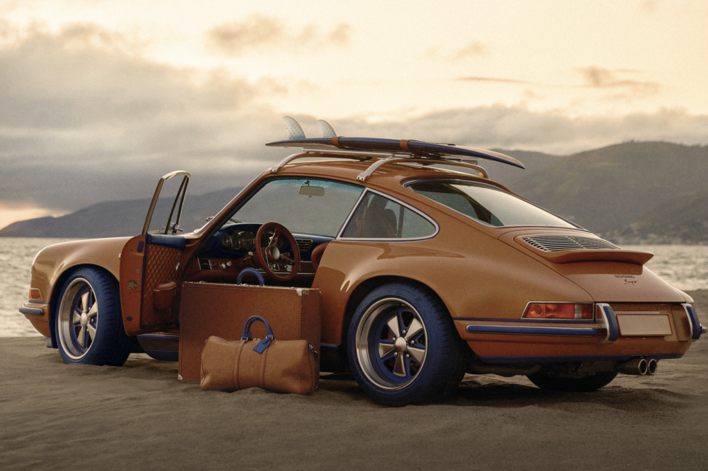 - Petróleo y Energía Orange vintage convertible with a surfboard on the roof, parked on a sandy beach with luggage in the foreground and ocean in the background.