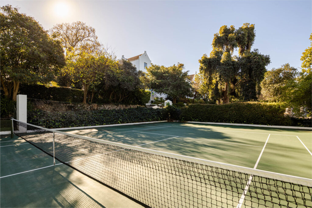 - Petróleo y Energía Outdoor tennis court with a net, surrounded by dense trees and hedges on a sunny day.