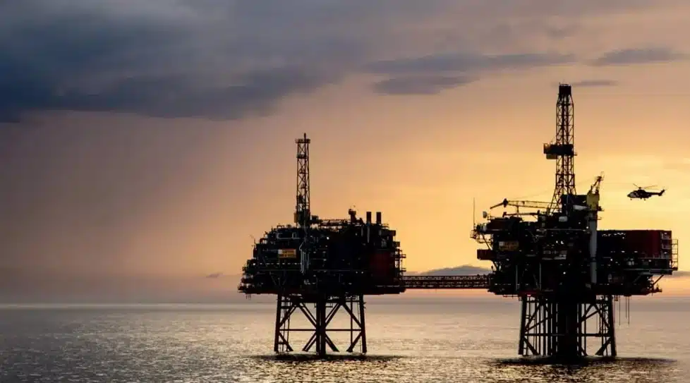 Two offshore oil rigs on stilts at sunset over calm water, with a helicopter nearby in the sky.