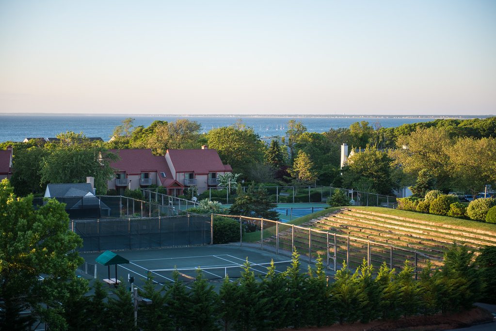 Nine Tennis Courts - Petróleo y Energía Coastal view of a park with tennis courts, bleachers, and red-roofed buildings in the foreground, ocean on the horizon.