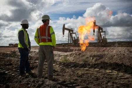 Two construction workers in hard hats and high‑visibility vests stand on a dirt oil field, watching a gas flare from an oil pump on site.