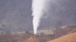 Vertical plume of white steam or vapor rising from a ground vent in a rural area, with leafless trees, fences, and rolling hills in the background.