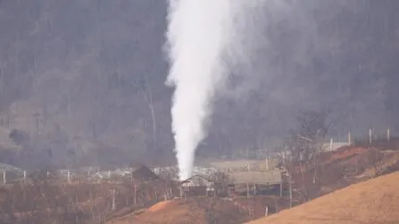 Vertical plume of white steam or vapor rising from a ground vent in a rural area, with leafless trees, fences, and rolling hills in the background.