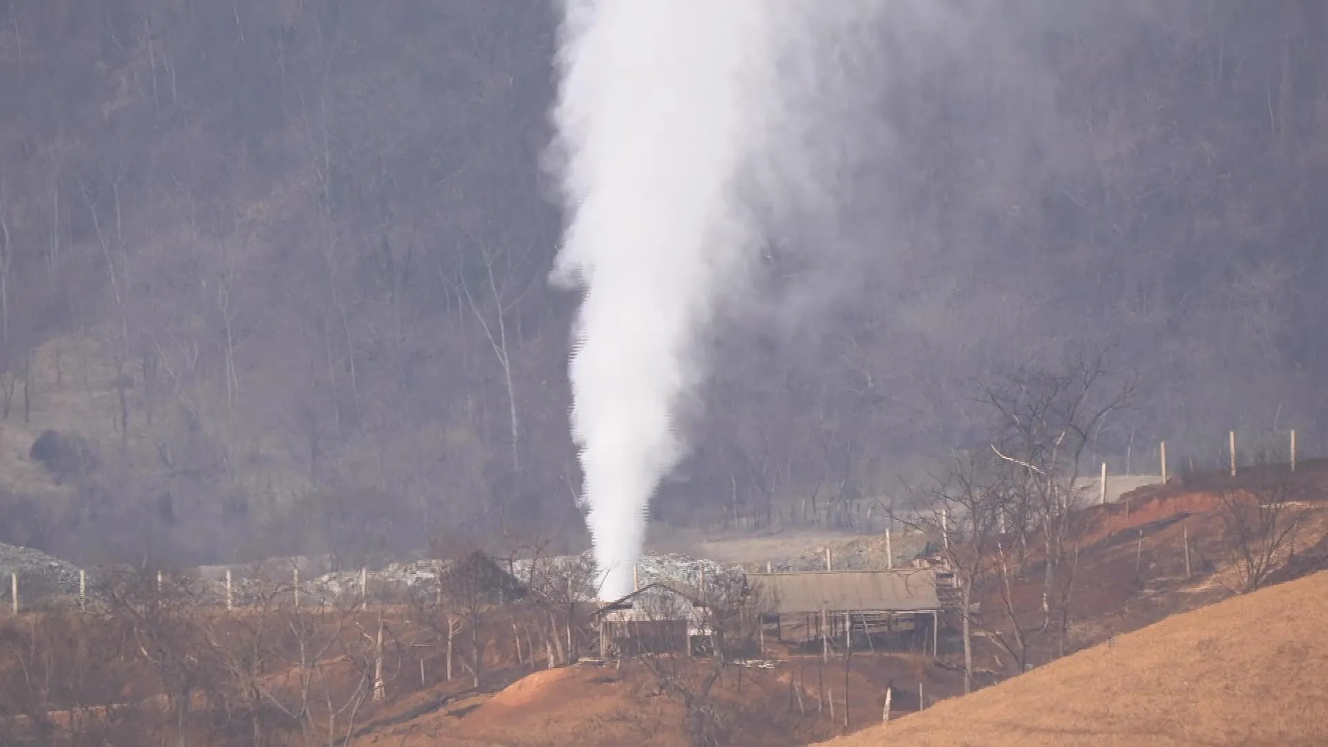 Vertical plume of white steam or vapor rising from a ground vent in a rural area, with leafless trees, fences, and rolling hills in the background.