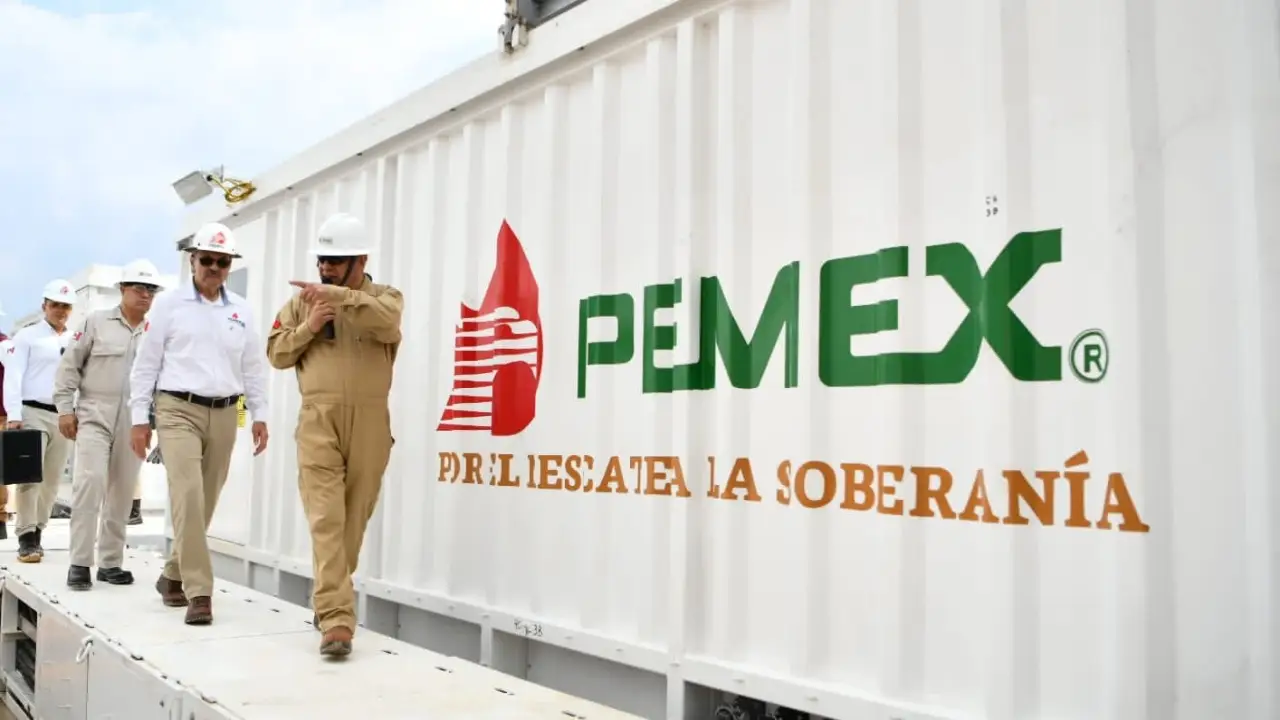 Group of engineers in hard hats walking along a white PEMEX container with red and green logo markings.